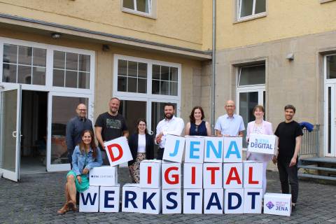 Neun Personen stehen vor einem Gebäude mit gestapelten Papierwürfeln, die jeweils einen Buchstaben abbilden und "Jena Digital Werkstadt" ergeben