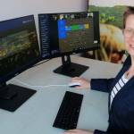 Manuela Meyer - CDO der Stadt Jena sitzt am Schreibtisch vor zwei Bildschirmen und schaut in die Kamera. Auf den Bildschrimen sieht man die Kick-Off Veranstaltung des Smart City Projekts.