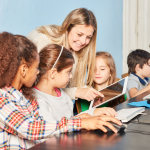 Kinder sitzen vor einem PC-Monitor und eine Lehrerin schaut lächelnd zu.