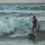 Mann surft auf einer Welle mit Smartphone in der Hand.