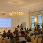 Zwei Personen die einen Vortrag gehalten haben an einem Pult, hinter ihnen eine Leinwand, mit einer Präsentation, vor Ihnen ein Publikum. Eine Person aus dem Publikum stellt eine Frage an die beiden Redner am Pult.