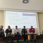 Personen sitzen auf dem Podium mit Mikro in der Hand, hinter sich eine PowerPoint