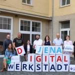 Neun Personen stehen vor einem Gebäude mit gestapelten Papierwürfeln, die jeweils einen Buchstaben abbilden und "Jena Digital Werkstadt" ergeben