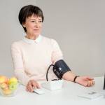 Zu sehen ist eine Frau die vor einem Computer sitzt und unter Anleitung ihren Blutdruck misst. Daneben steht eine Schale mit Äpfeln.