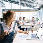 Frau arbeitet am Laptop im Büro, im Hintergrund unterhalten sich Menschen.