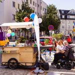 Ein mobiler Infostand mit bunten Luftballons und Pflanzen steht auf einem Gehweg in einer Stadt, während eine Frau im Gespräch mit einem Mann im Elektrorollstuhl ist. Im Hintergrund sind parkende Autos, Wohnhäuser und Passanten zu sehen.