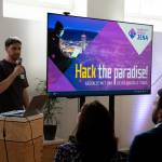 Domenique Dölz, Teilprojektleiter Handlungsfeld "Wirtschaft und Wissenschaft", steht mit einem Mikrofon und Bildschirm mit der Aufschrift "Hack the paradise! Gestalte mit uns deine digitale Stadt der Zukunft" vor einem Publikum