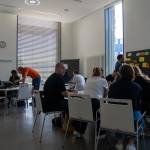 Viele Menschen sitzen in einem Raum in Gruppen an Tischen und arbeiten gemeinsam, im Hintergrund stehen Pinnwände mit verschiedenen Zetteln