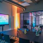 Ein Raum mit einer Glastür und zusammengeschobenen Tischen auf denen Getränke und Blumen stehen; ein Bildschirm auf einem Gestell mit dem Aufzug "Jena Digital - Opening Week 2024"