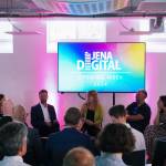 Ein Raum mit einem Bildschirm auf dem "Jena Digital - Opening Week 2024" steht und davor in einem Halbkreis sitzen 7 Personen, eine davon mit einem Mikrofon, dahinter sitzen Menschen in Stuhlreihen und schauen auf den Bildschirm 