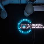 Eine Wand mit einer Leuchtreklame, die aus einem blauen Kreis mit weißer Schrift "JEDI - Jena Digital Innovation Hub", die den Kreis durchbricht, besteht 