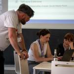 Ein Mann lehnt über einen Stuhl und schaut zu zwei Frauen die am Tisch sitzen und sich besprechen