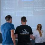 Zwei Männer und eine Frau stehen mit dem Rücken zur Kamera und schauen auf ein Computer-Skript, welches an der Wand über einen Beamer zu sehen ist.