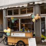 Holz-Anhänger für ein Lastenfahrrad mit der Aufschrift „ProbierMOBIL“ vor einem Ladenlokal, das mit gelb-grünen Luftballons geschmückt ist.