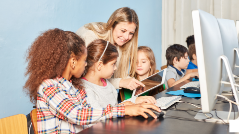Kinder sitzen vor einem PC-Monitor und eine Lehrerin schaut lächelnd zu.