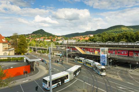 Foto zeigt Ausschnitt vom Busbahnhof Jena und Paradiesbahnhof. In der Mitte fahren Bus und Straßenbahn, im Hintergrund Blick auf den Jenzig.