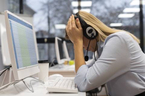 Frau mit Kopfhörern sitzt vor einem Monitor und hat müde den Kopf in die Hände gestützt.