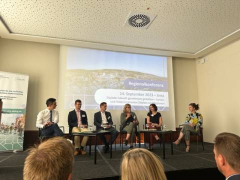 Podium mit sechs Personen vor einer Leinwand, vor ihnen das Publikum