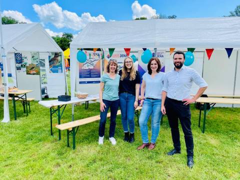 v.l.: Heike Stötzner, Vanessa Huber, Dorothea Prell und Benjamin Koppe stehen vor dem Smart City Stand bei "Dein Tag im Paradies" 2024. . Im Hintergrund sieht man ein Zelt mit Plakaten des Projekts "5G-Verkehrsvernetznung" und "Smart City Projekt".