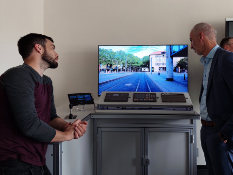 In der Mitte der neue Fahrerstand beim Nahverkehr zum Trainieren des Fahrerassistenzsystem. Daneben stehen rechts und links 2 Männer, die sich auf dem Computer in der Mitte das neue System anschauen.