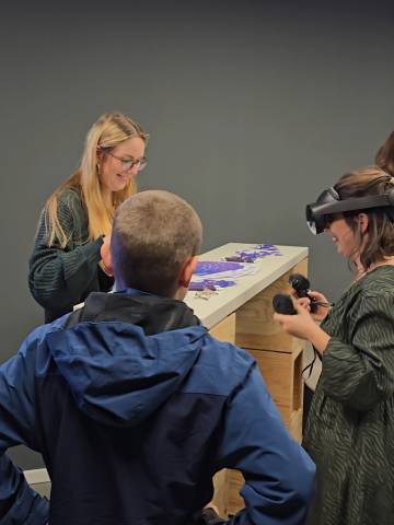 Drei Personen in einem Raum, eine davon trägt eine sogenannte Virtual Reality Brille.