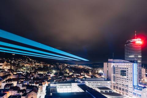 Die Gebäude in Jena erstrahlen im Dunkeln, am Himmel ist ein großer Laserstrahl.