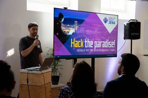 Domenique Dölz, Teilprojektleiter Handlungsfeld "Wirtschaft und Wissenschaft", steht mit einem Mikrofon und Bildschirm mit der Aufschrift "Hack the paradise! Gestalte mit uns deine digitale Stadt der Zukunft" vor einem Publikum