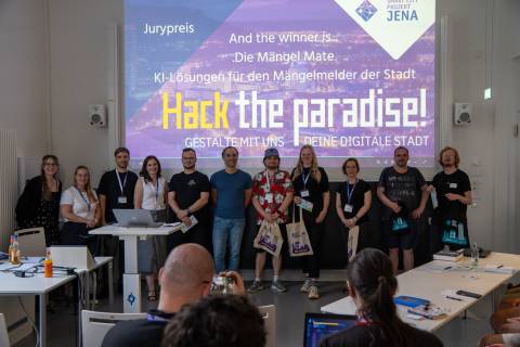 Vor einer Leinwand mit der Schrift "Hack the paradise! Gestalte mit uns deine digitale Stadt" stehen mehrere Menschen in einer Reihe mit Smart City Beuteln in der Hand vor einem Publikum
