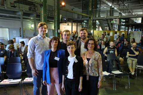 Mehrere Personen stehen in einer Halle und schauen in die Kamera, im Hintergrund sitzen Menschen.