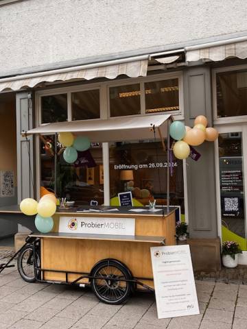 Holz-Anhänger für ein Lastenfahrrad mit der Aufschrift „ProbierMOBIL“ vor einem Ladenlokal, das mit gelb-grünen Luftballons geschmückt ist.