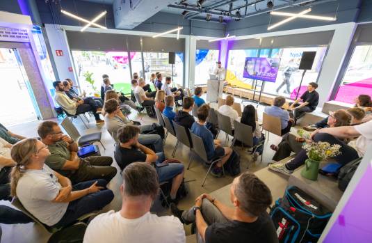 Mann steht am Pult und spricht vor einem Publikum, auf einem Bildschirm läuft eine Präsentation zum Hackathon "Hack the paradise!"