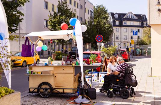 Ein mobiler Infostand mit bunten Luftballons und Pflanzen steht auf einem Gehweg in einer Stadt, während eine Frau im Gespräch mit einem Mann im Elektrorollstuhl ist. Im Hintergrund sind parkende Autos, Wohnhäuser und Passanten zu sehen.