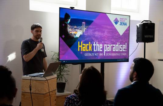 Domenique Dölz, Teilprojektleiter Handlungsfeld "Wirtschaft und Wissenschaft", steht mit einem Mikrofon und Bildschirm mit der Aufschrift "Hack the paradise! Gestalte mit uns deine digitale Stadt der Zukunft" vor einem Publikum