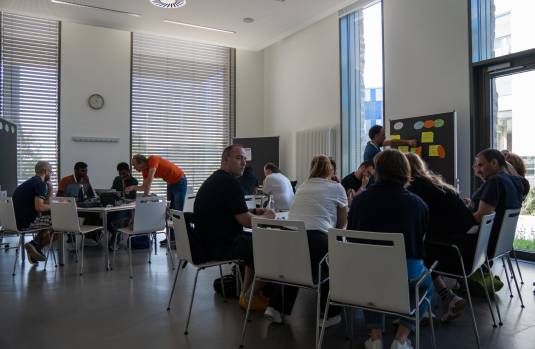 Viele Menschen sitzen in einem Raum in Gruppen an Tischen und arbeiten gemeinsam, im Hintergrund stehen Pinnwände mit verschiedenen Zetteln