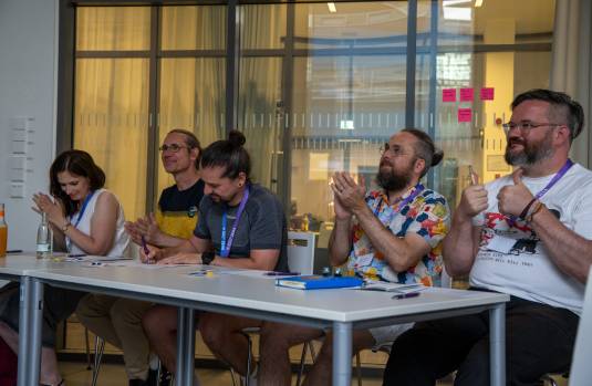 Eine Frau und 4 Männer sitzen an einem Tisch, klatschen in die Hände und sind erfreut.