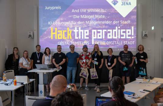 Vor einer Leinwand mit der Schrift "Hack the paradise! Gestalte mit uns deine digitale Stadt" stehen mehrere Menschen in einer Reihe mit Smart City Beuteln in der Hand vor einem Publikum