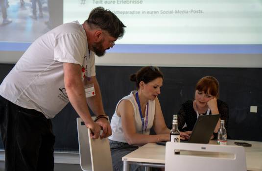 Ein Mann lehnt über einen Stuhl und schaut zu zwei Frauen die am Tisch sitzen und sich besprechen