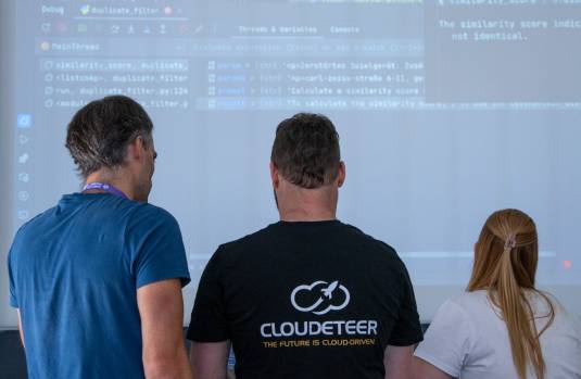 Zwei Männer und eine Frau stehen mit dem Rücken zur Kamera und schauen auf ein Computer-Skript, welches an der Wand über einen Beamer zu sehen ist.