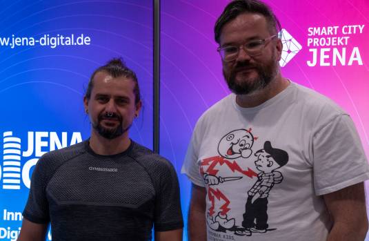Zwei Männer stehen vor einer Leuchtreklame des Jena Digital Vereins und lächeln in die Kamera.