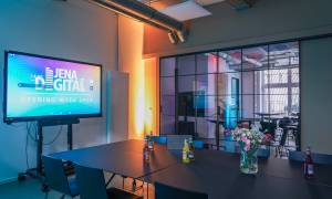 Ein Raum mit einer Glastür und zusammengeschobenen Tischen auf denen Getränke und Blumen stehen; ein Bildschirm auf einem Gestell mit dem Aufzug "Jena Digital - Opening Week 2024"