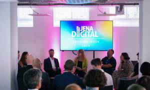 Ein Raum mit einem Bildschirm auf dem "Jena Digital - Opening Week 2024" steht und davor in einem Halbkreis sitzen 7 Personen, eine davon mit einem Mikrofon, dahinter sitzen Menschen in Stuhlreihen und schauen auf den Bildschirm 