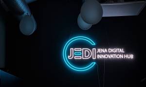 Eine Wand mit einer Leuchtreklame, die aus einem blauen Kreis mit weißer Schrift "JEDI - Jena Digital Innovation Hub", die den Kreis durchbricht, besteht 