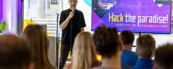 Moderator spricht vor Publikum mit einer Präsentation zum Hackathon "Hack the paradise!"