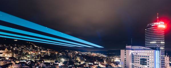 Die Gebäude in Jena erstrahlen im Dunkeln, am Himmel ist ein großer Laserstrahl.