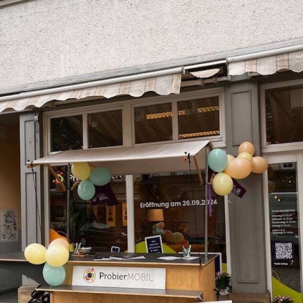 Holz-Anhänger für ein Lastenfahrrad mit der Aufschrift „ProbierMOBIL“ vor einem Ladenlokal, das mit gelb-grünen Luftballons geschmückt ist.