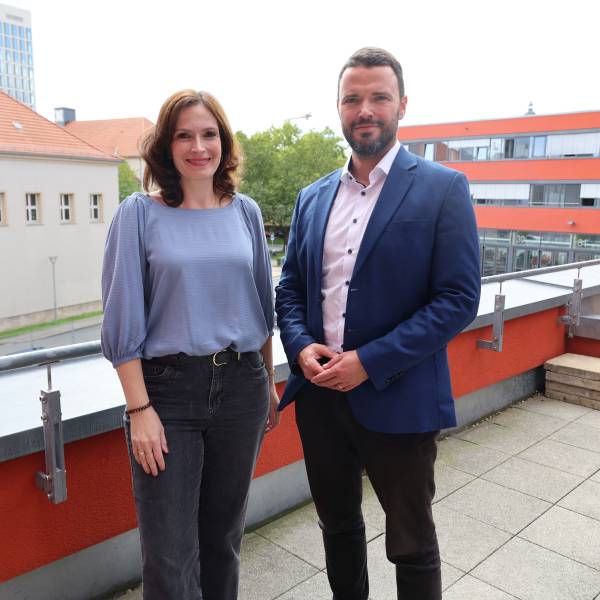 Mann und Frau stehen auf einer Dachterrasse mit Jena im Hintergrund und lächeln in die Kamera.
