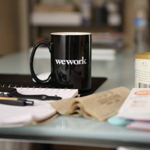 Blick auf einen Schreibtisch, auf dem eine Kaffeetasse, Stifte, Uhr und Telle liegen. Auf der Tasse steht "we work".