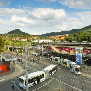 Foto zeigt Ausschnitt vom Busbahnhof Jena und Paradiesbahnhof. In der Mitte fahren Bus und Straßenbahn, im Hintergrund Blick auf den Jenzig.