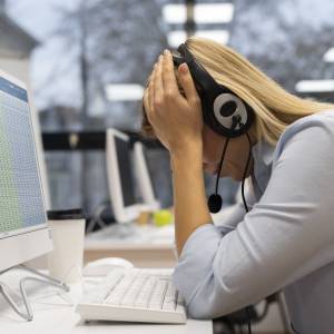 Frau mit Kopfhörern sitzt vor einem Monitor und hat müde den Kopf in die Hände gestützt.