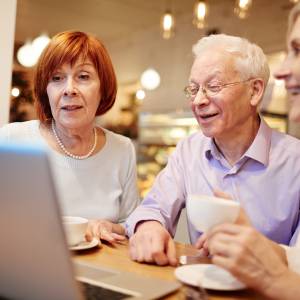 Drei ältere Personen sitzen lächelnd vor einem Laptop mit einer Tasse Kaffee in der Hand. in 
